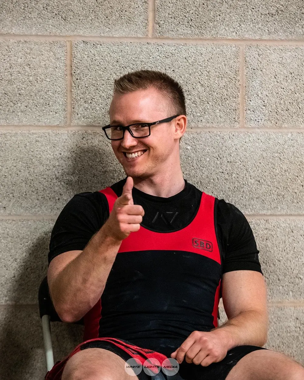 Andrew sitting against a breeze-block wall, smiling and pointing at the camera