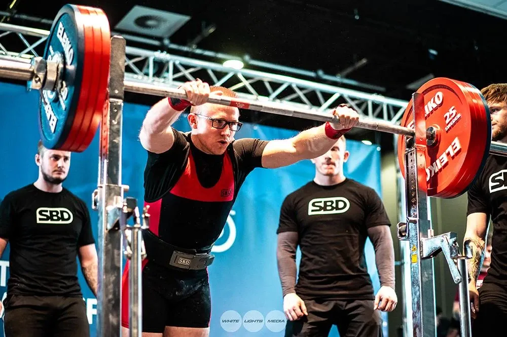 Andrew looking serious as he grips the squat bar, with two spotters behind