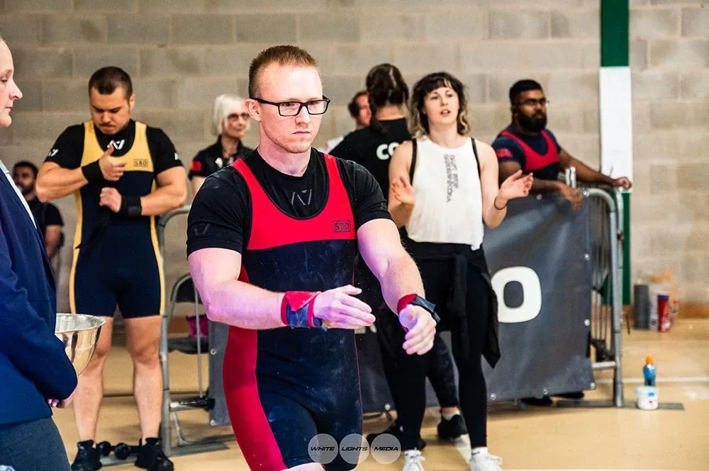 Andrew walking to the platform for his second attempt, looking serious, with other lifters and coaches behind him