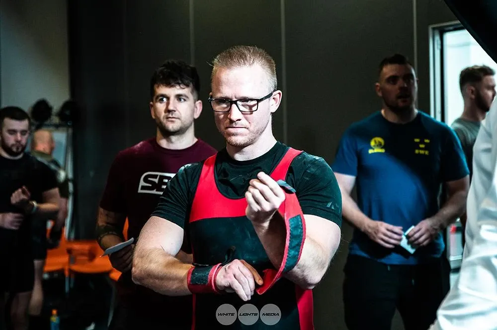 Andrew preparing his wrist wraps as he gets ready to lift, with other lifters and coaches behind him