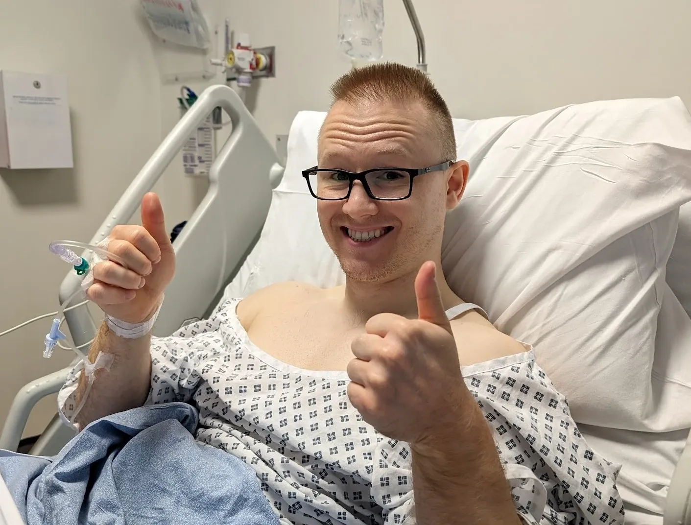 Andrew in a hospital gown sitting up in bed, grinning and giving two thumbs up, with a cannula in his hand