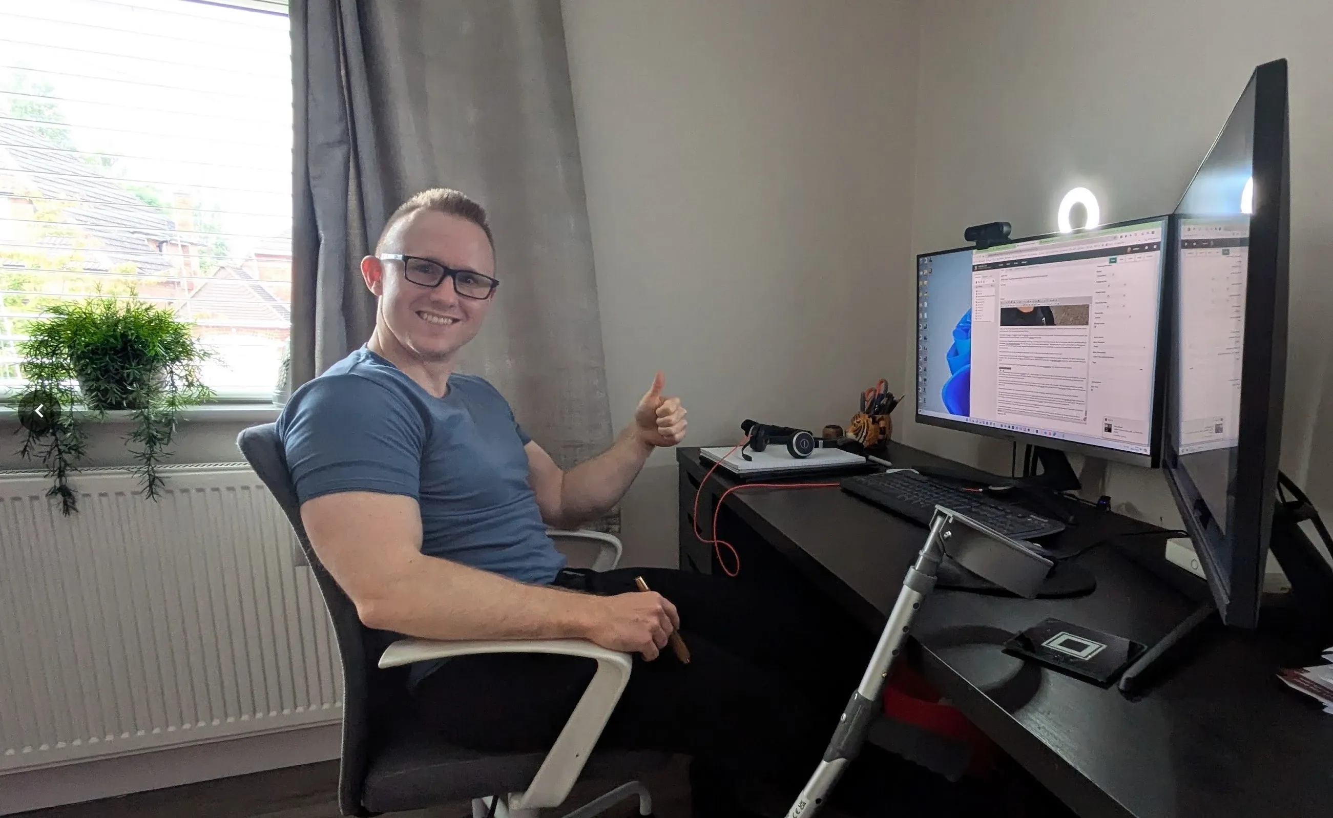 Andrew smiling and giving a thumbs up at his desk in front of two monitors, with a crutch leaning against the desk