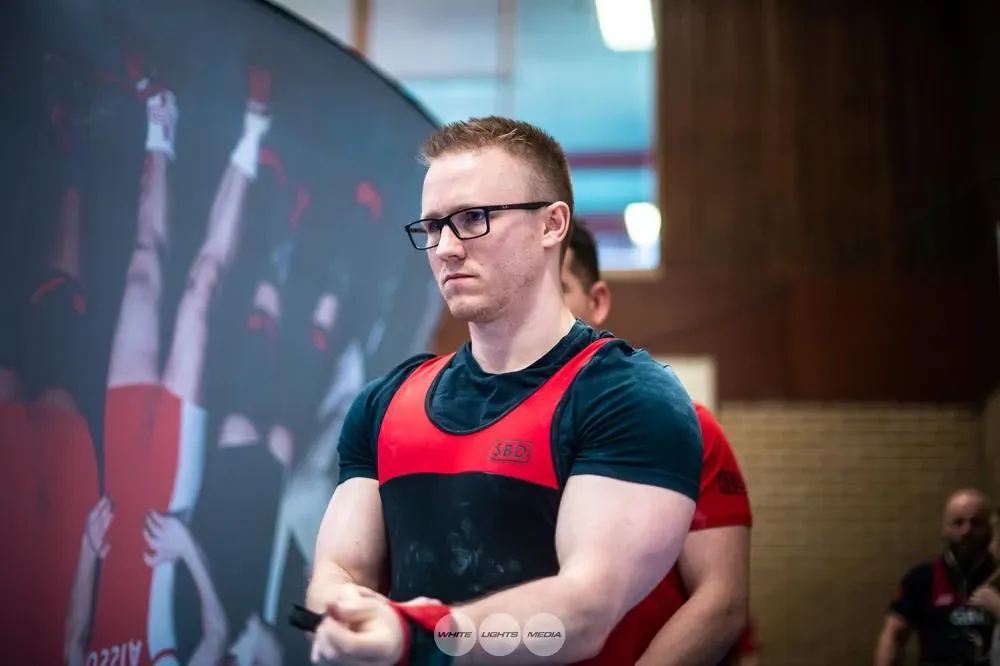 Andrew waiting to bench press, looking focused as he adjusts his wrist wrap