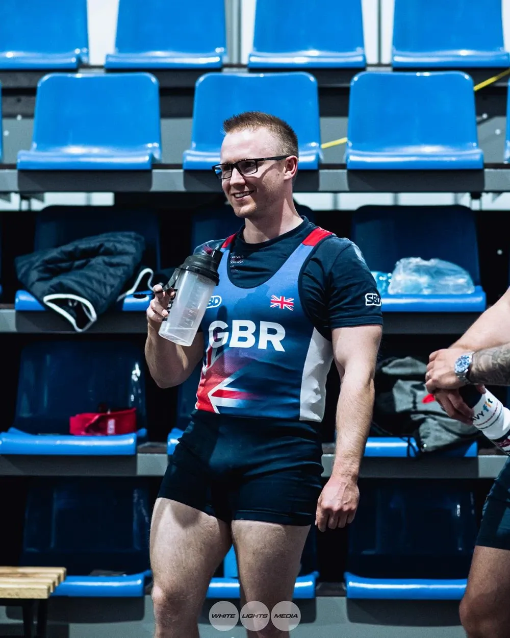 Andrew in the Great Britain singlet, smiling and drinking from a shaker bottle in the stands as he gets in the zone