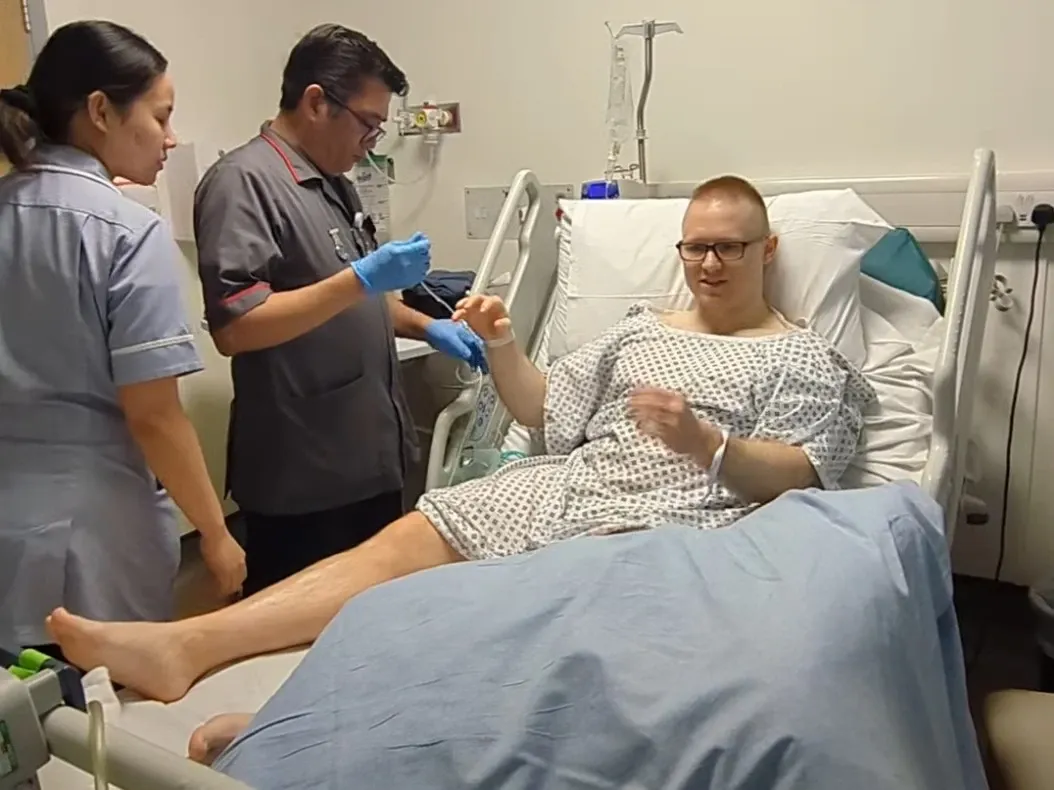 Andrew sitting up in a hospital bed in a gown while two members of staff attend to him