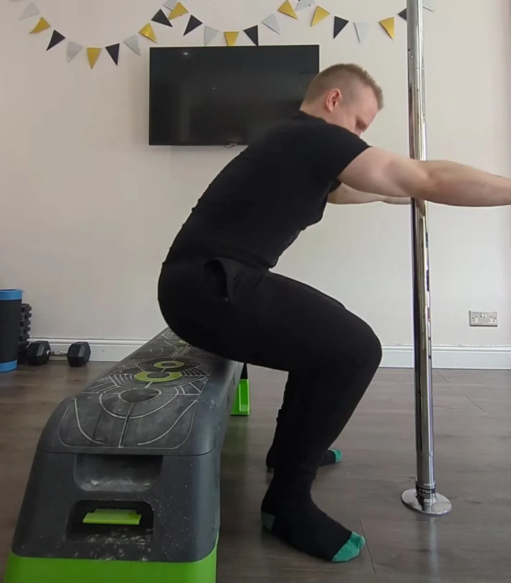 Andrew squatting to a low step bench at home with his arms held out in front, not holding the pole