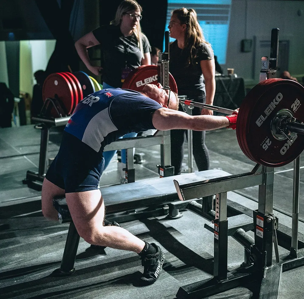 Andrew in a Great Britain singlet arching on the bench as he reaches for the bar, with loaders and a referee watching
