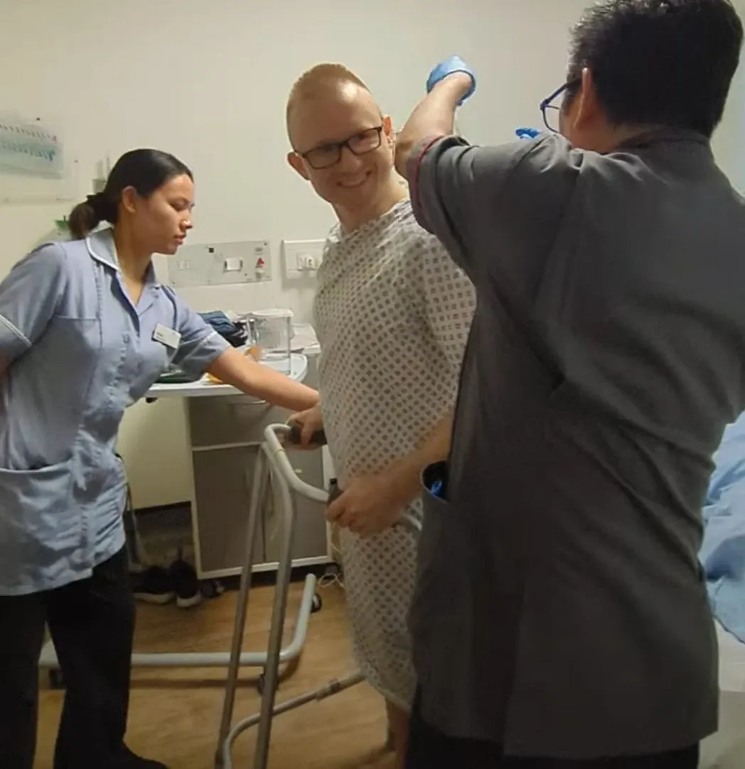 Andrew standing with a walking frame in his hospital room, smiling, with two members of staff beside him