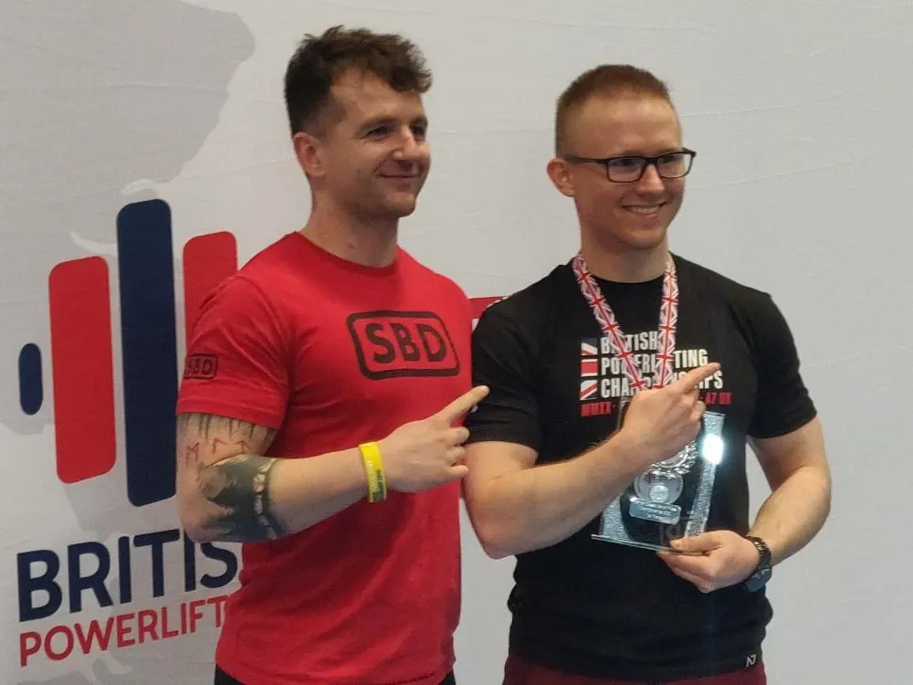 Andrew and Luke Rogers pointing at the trophy to celebrate the win, in front of a British Powerlifting backdrop
