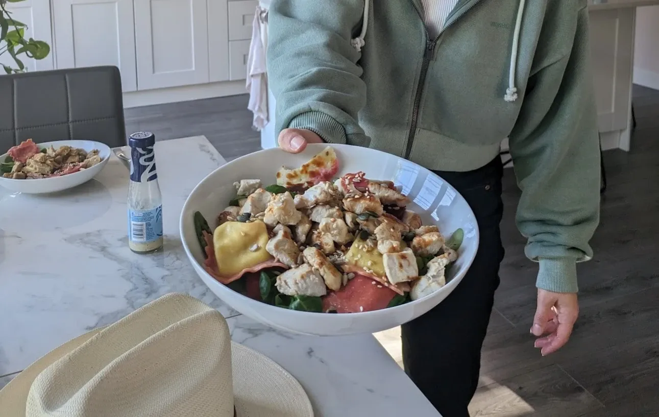 A large bowl of salad topped with chunks of chicken, pasta parcels, halloumi and seeds, held out over a kitchen table