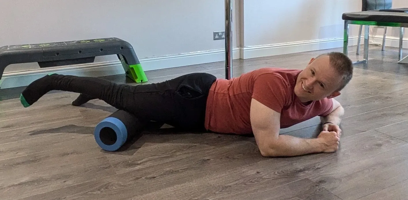 Andrew lying on his side on a wooden floor, smiling at the camera, with a foam roller under his inner thigh