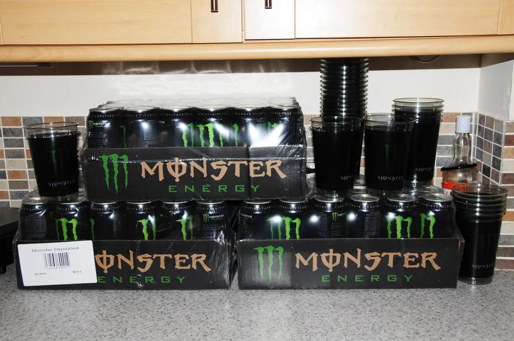 Three shrink-wrapped trays of Monster Energy cans and stacks of Monster cups on a kitchen worktop