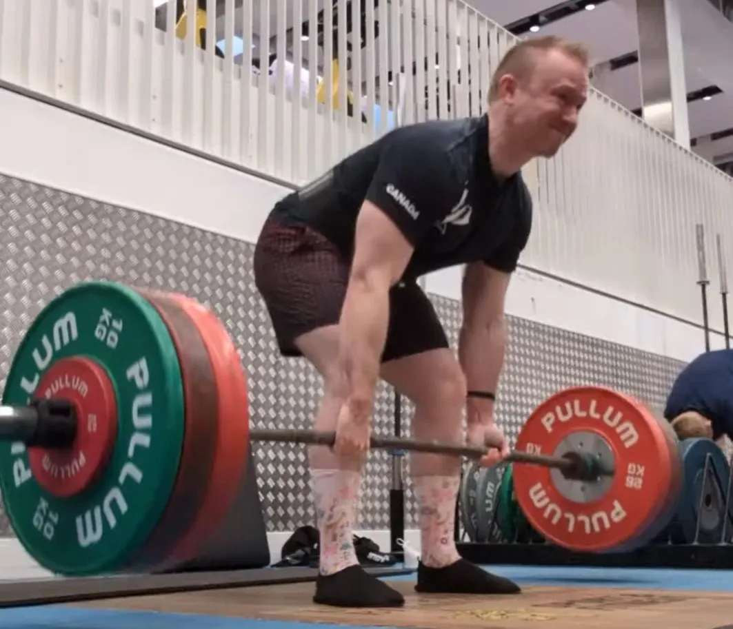 Andrew at the start of a deadlift, gripping a barbell loaded with competition plates in a gym