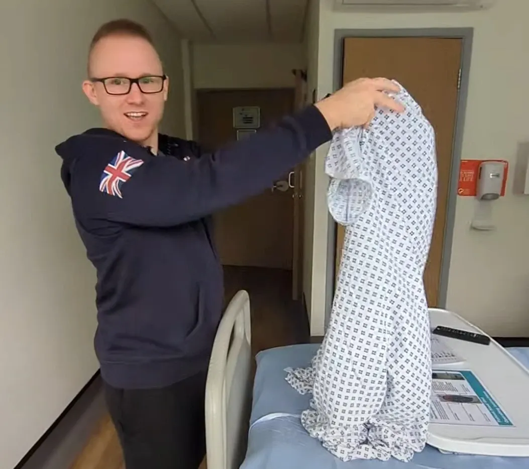 Andrew in a hoodie holding up a hospital gown beside a hospital bed