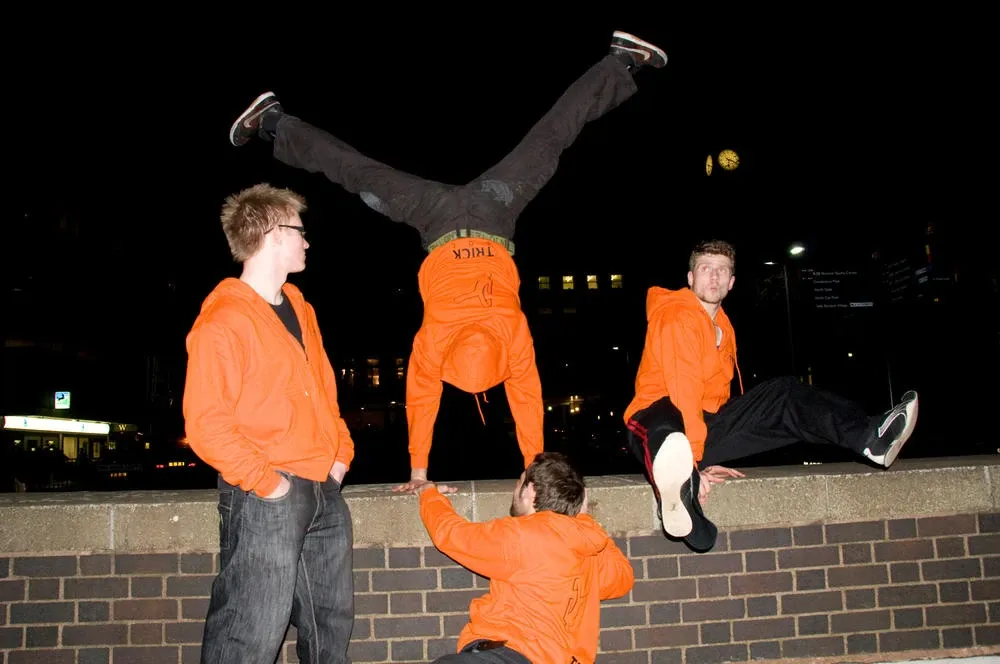 Andrew, Ed, Andy and Andy in orange Trick Soc hoodies by a brick wall at night, one of them in a handstand on top of the wall