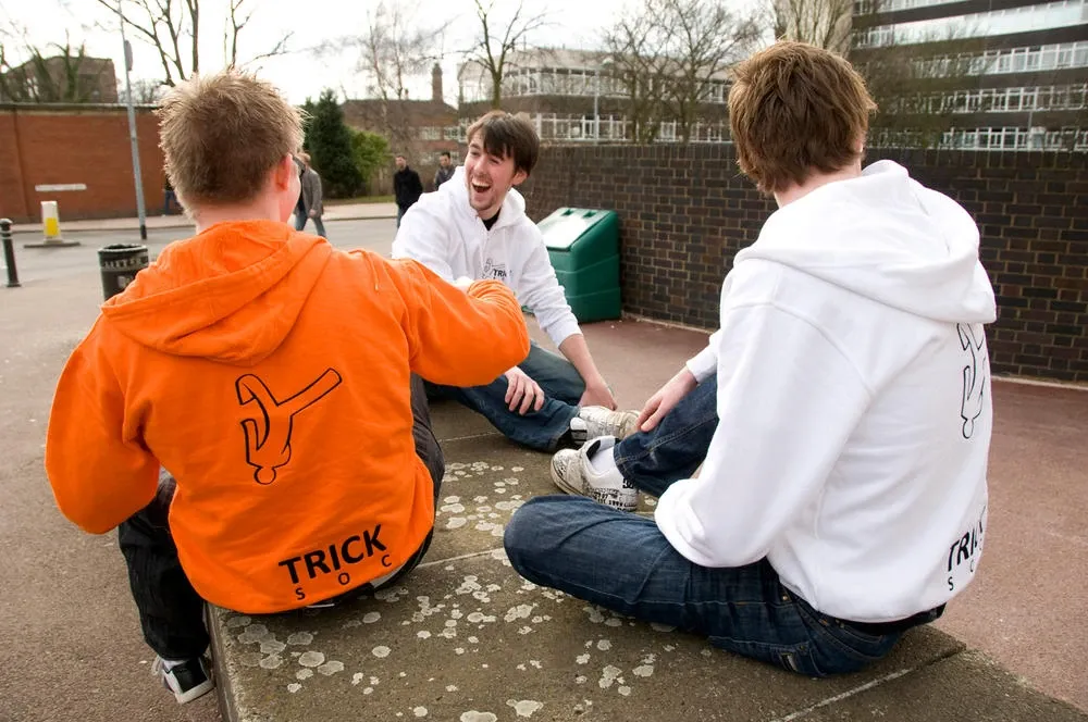 Andrew Ward, Mike Williams and Jared Cole sitting on a low wall on campus in orange and white Trick Soc hoodies