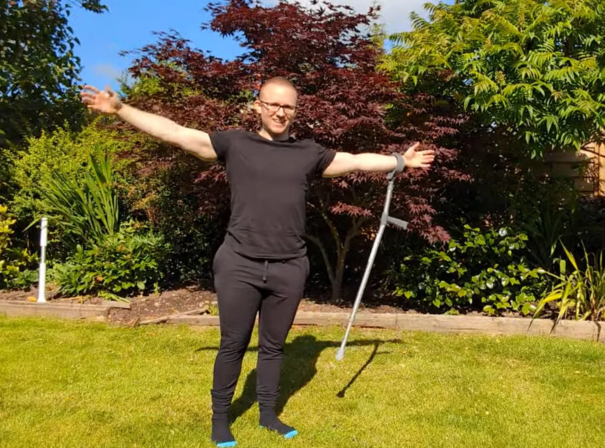 Andrew standing on his lawn with his arms spread wide and a crutch hanging from one wrist