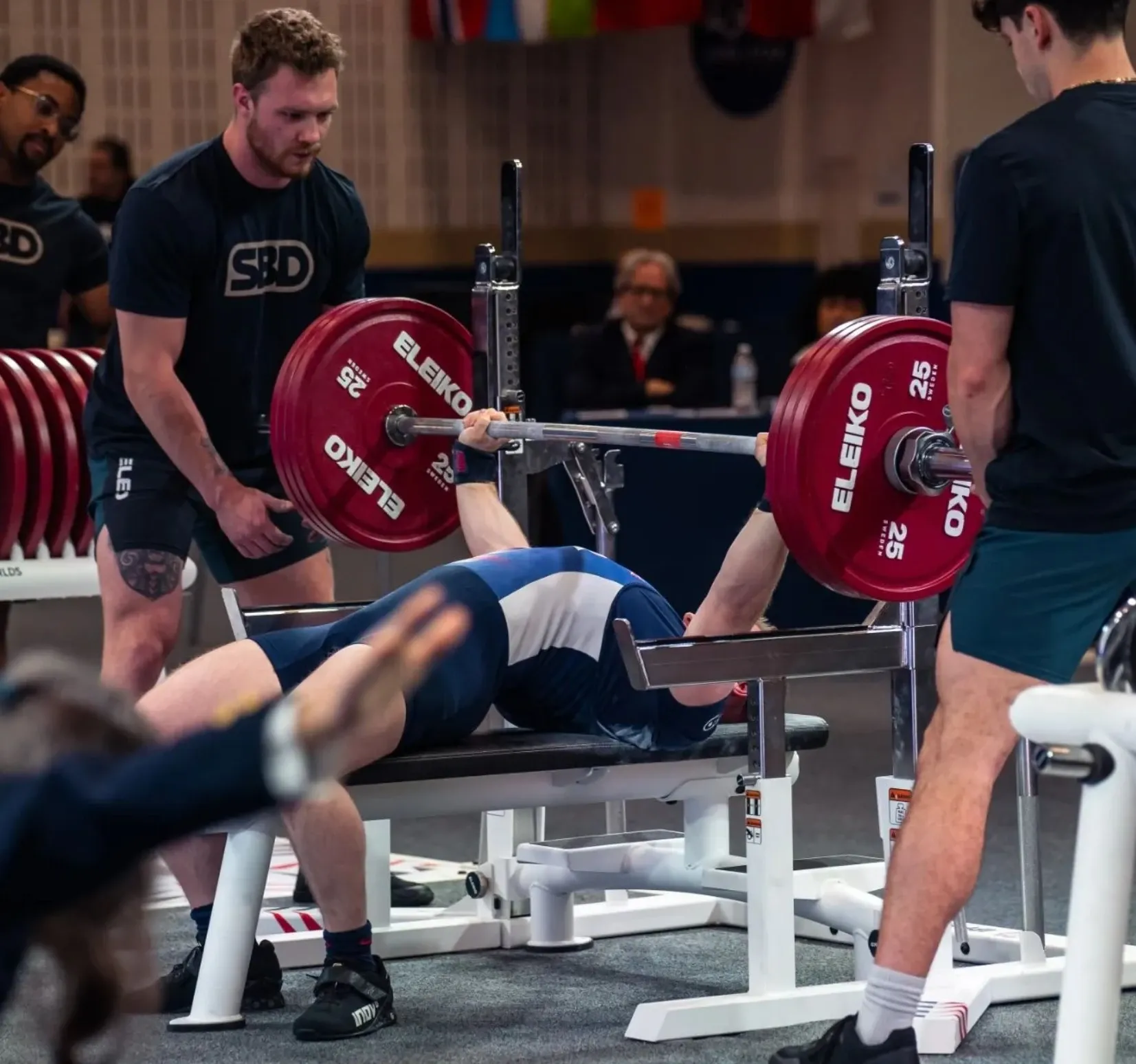 Andrew lowering a loaded barbell to his chest on the competition bench at the IPF World Championships, with spotters either side